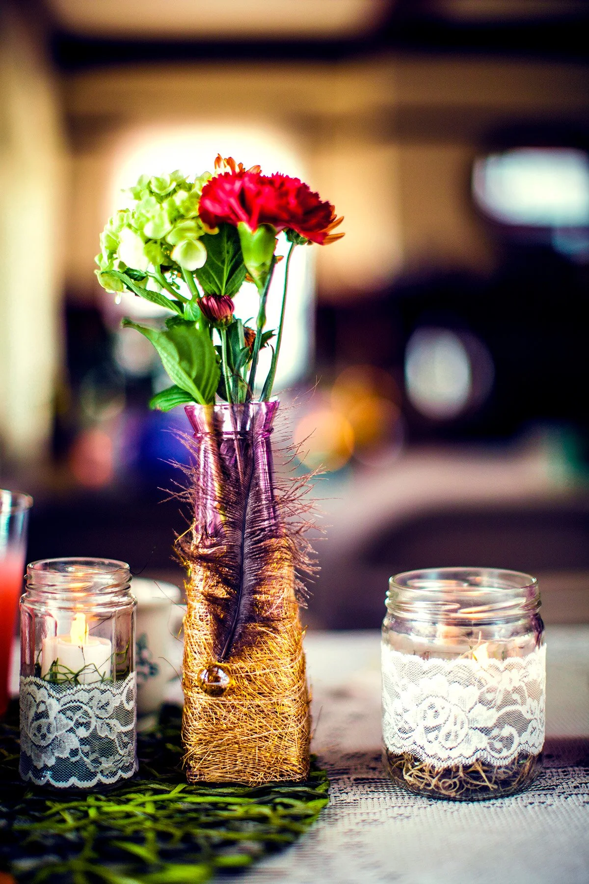 A small bouquet of flowers, including green and red blooms, arranged in a purple glass vase wrapped in a brown woven material, placed on a table with two candles in glass jars on either side.