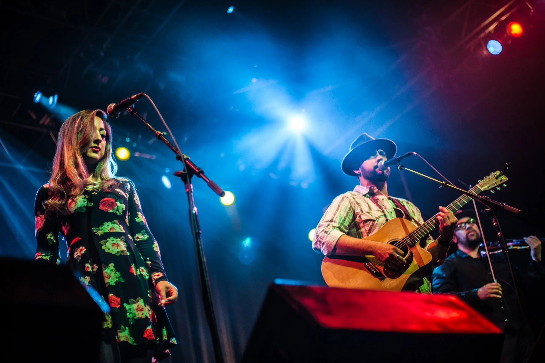 Musicians performing on stage under colorful lights, including a woman in a floral dress singing into a microphone and a man wearing a hat playing an acoustic guitar.