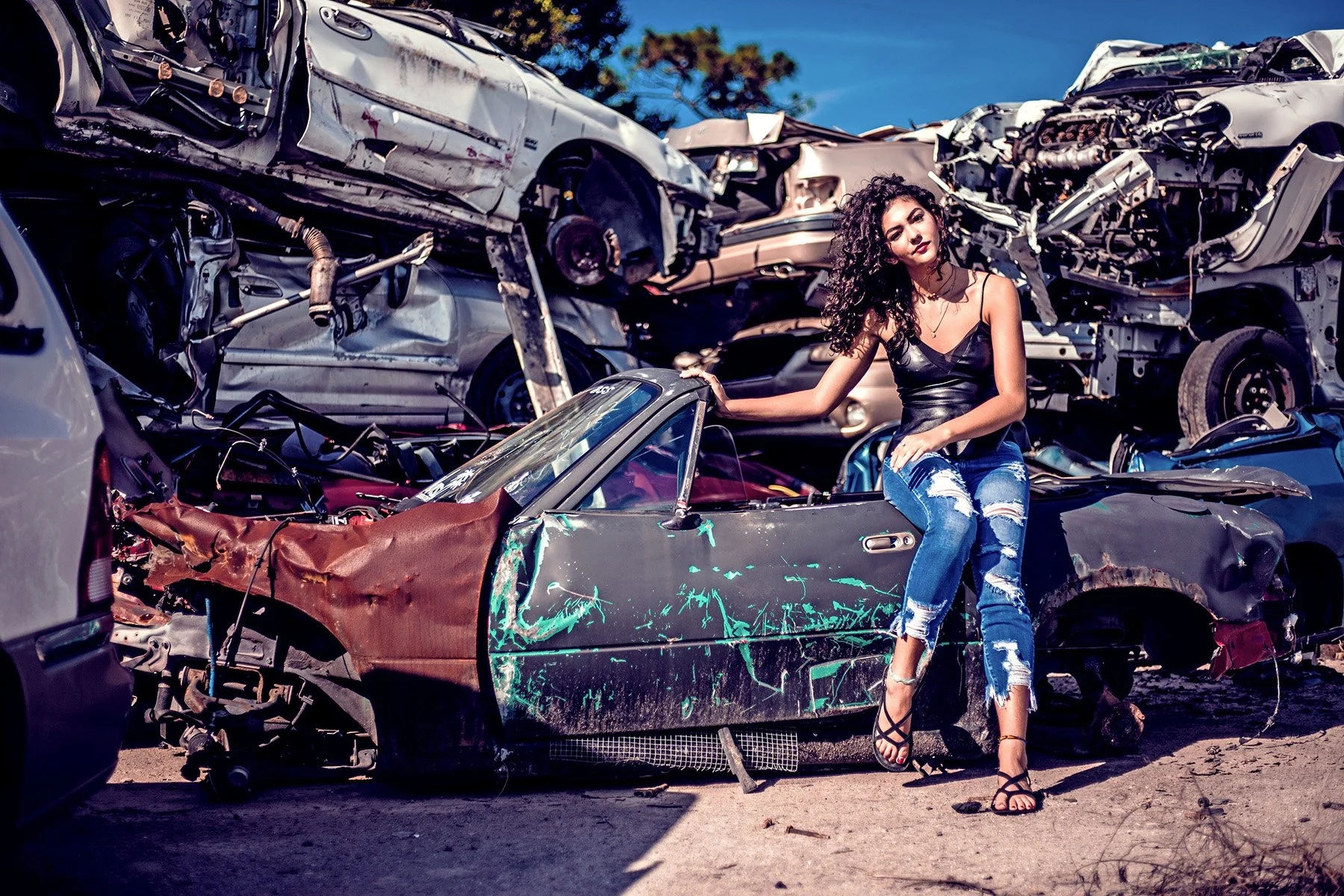 A woman with curly hair, wearing a black leather top and ripped jeans, sitting on a crushed sports car at a junkyard filled with wrecked cars.