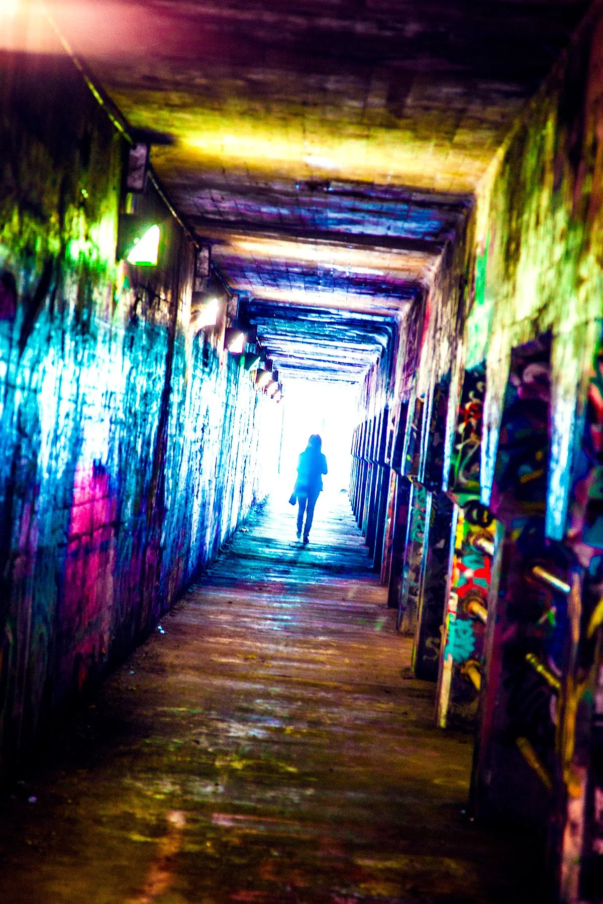 A person walking through a tunnel with graffiti-covered walls illuminated by colorful lights, leading towards a bright light at the end.
