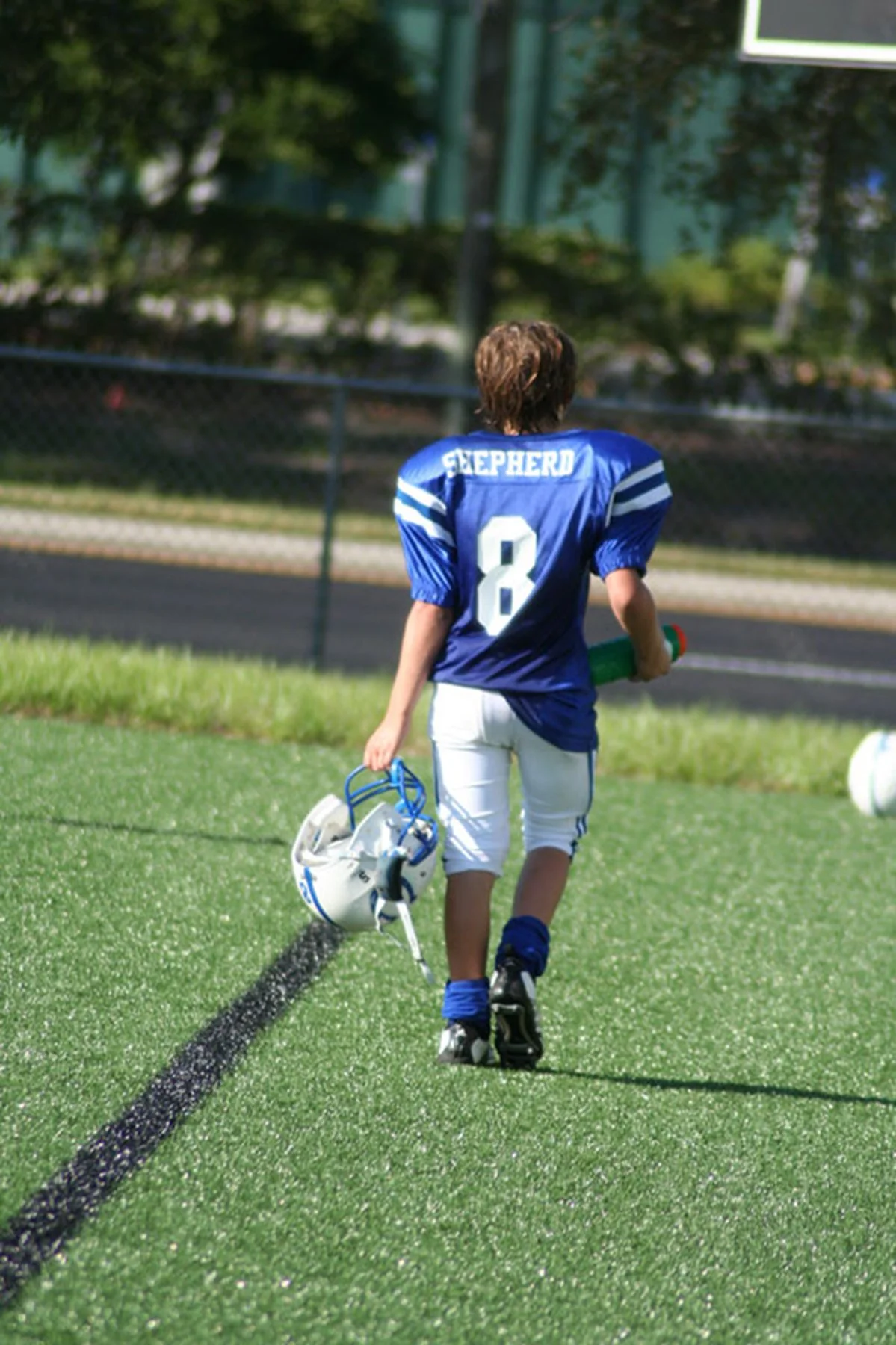 A young football player walking on a field holding a helmet and water bottle, wearing a blue jersey with the number 8 and the word 'SHEPHERD' on the back.