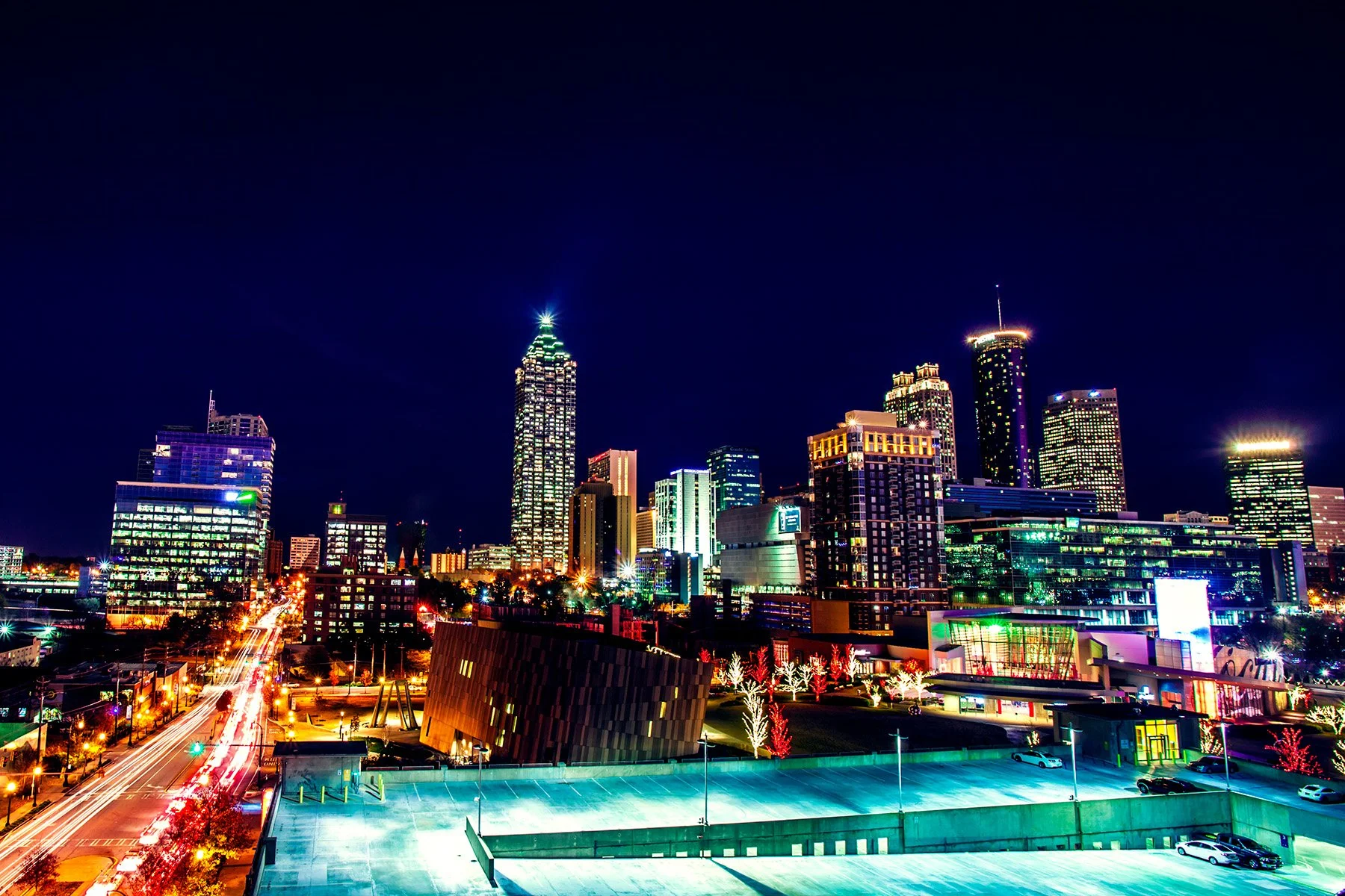 Night skyline of a city with tall illuminated skyscrapers and colorful lights reflecting in the buildings, with streets below showing light trails from moving vehicles.