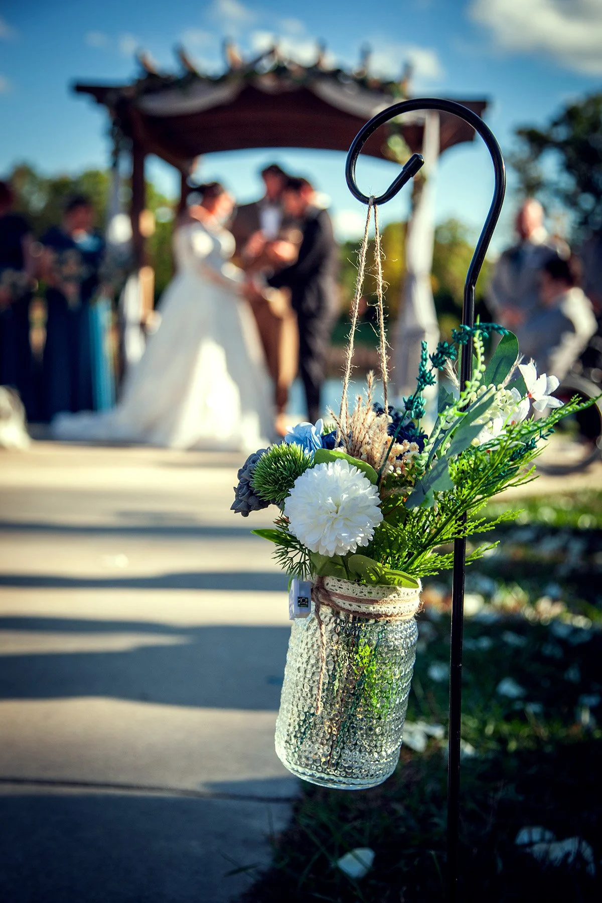 Close-up of a decorative green and white floral arrangement in a bejeweled silver jar hanging from a black hook, with a blurred outdoor wedding ceremony in the background
