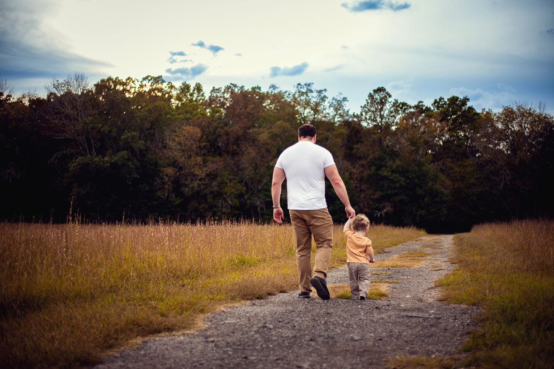 A man and a young child walking hand in hand along a dirt path in a grassy field with trees in the background during daytime.