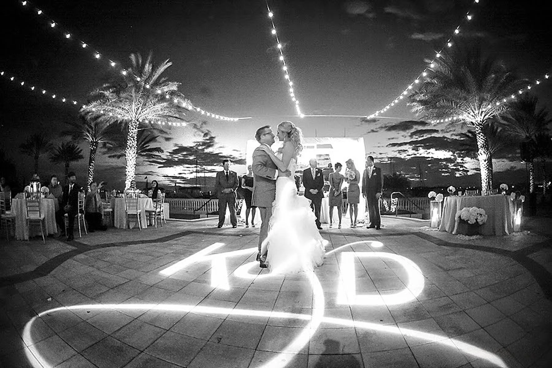A couple in wedding attire dancing at an outdoor evening wedding reception, illuminated by string lights and palm trees with a sunset sky in the background.