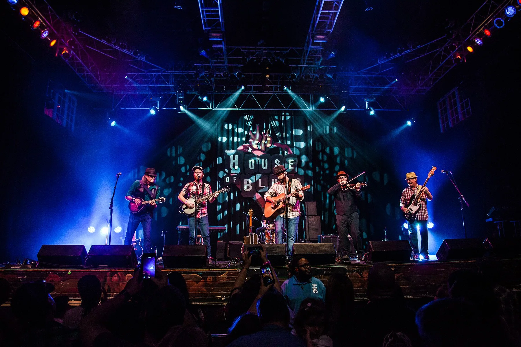 Musicians performing on stage at the House of Blues, illuminated by colorful stage lights, with audience members taking photos.