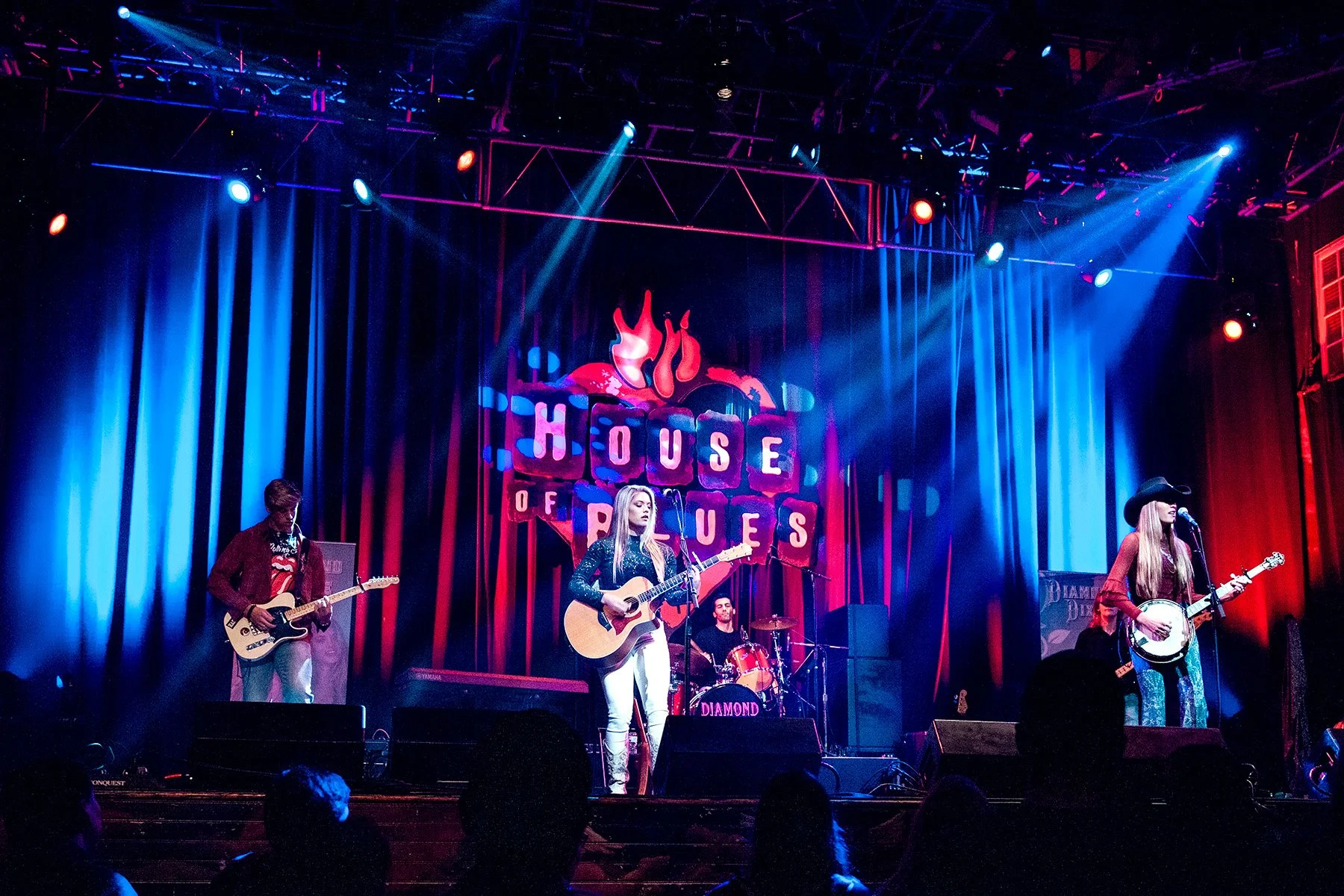 A live music performance on stage at the House of Blues, featuring a band with female singers, a guitarist, a bassist, and a drummer. The stage is decorated with colorful lights and a backdrop displaying the House of Blues logo.