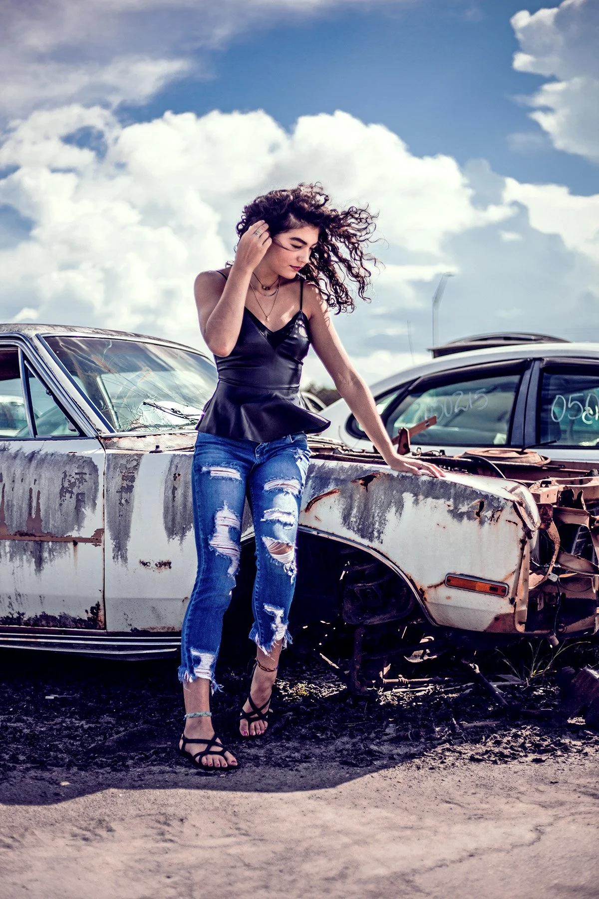 A young woman with dark, curly hair standing next to a rusty, old car in a junkyard. She is wearing a black sleeveless top, ripped jeans, and high heels, with her hand on her head and looking down.