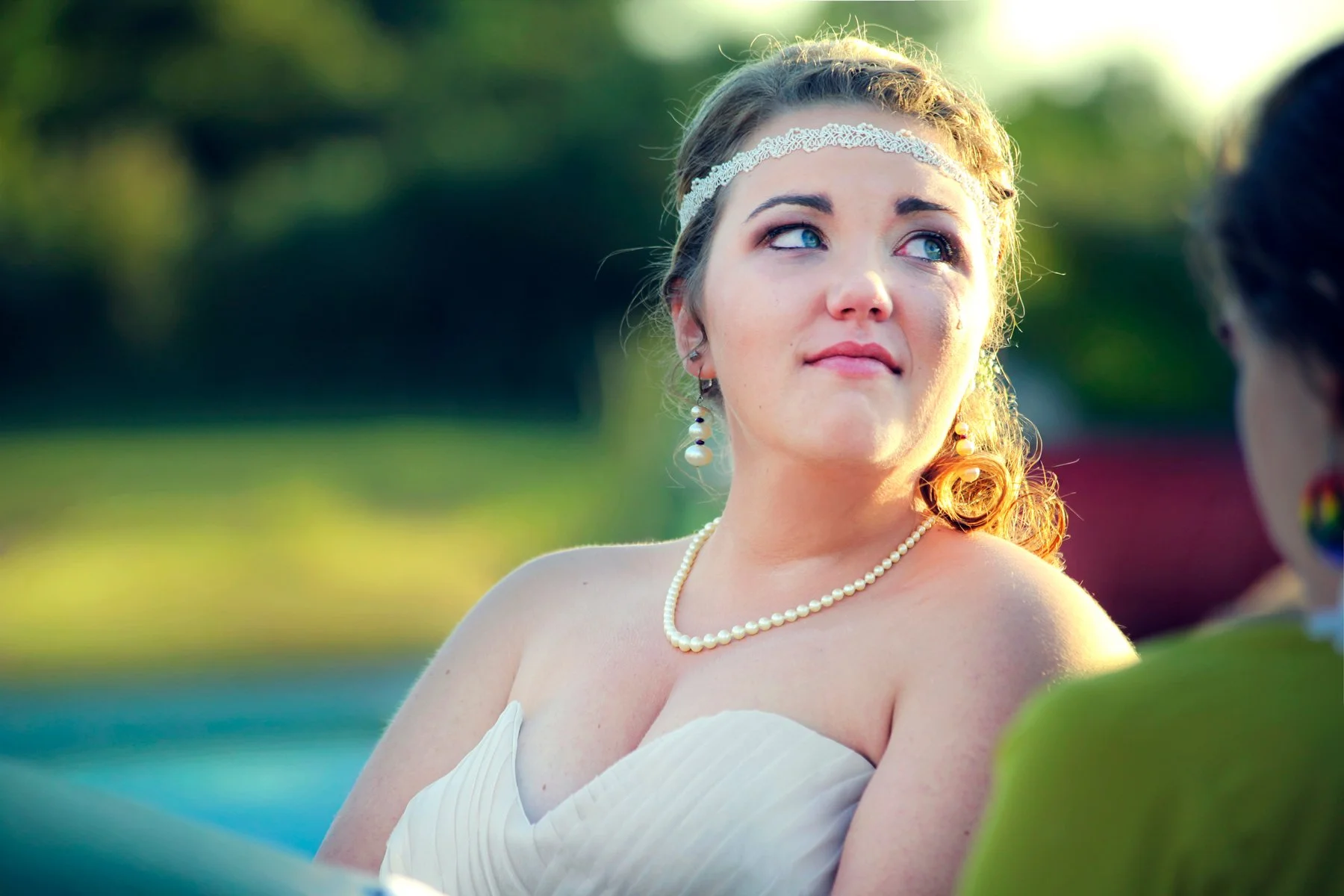 A woman in a white strapless dress, pearl necklace, and pearl earrings looks to her right with a tear running down her face during an outdoor event, with sunlight and a blurry green background.