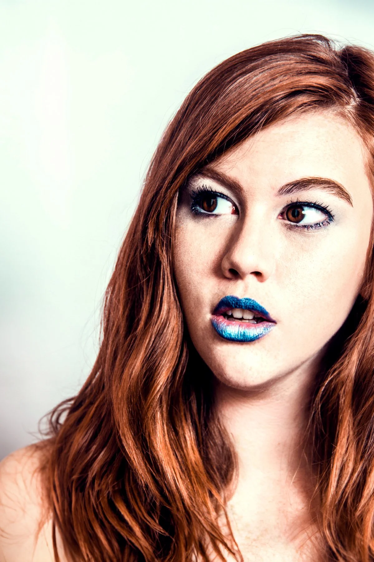 Close-up of a woman with red hair, brown eyes, bold blue lipstick, and makeup, looking to the side against a plain light background.