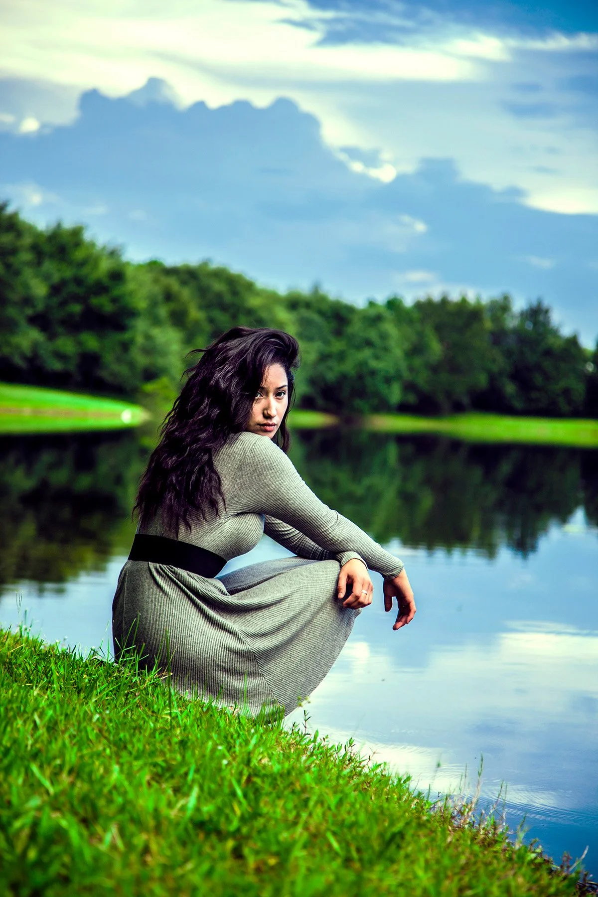 A woman with long dark wavy hair sitting by a calm lake surrounded by lush green trees under a partly cloudy sky.