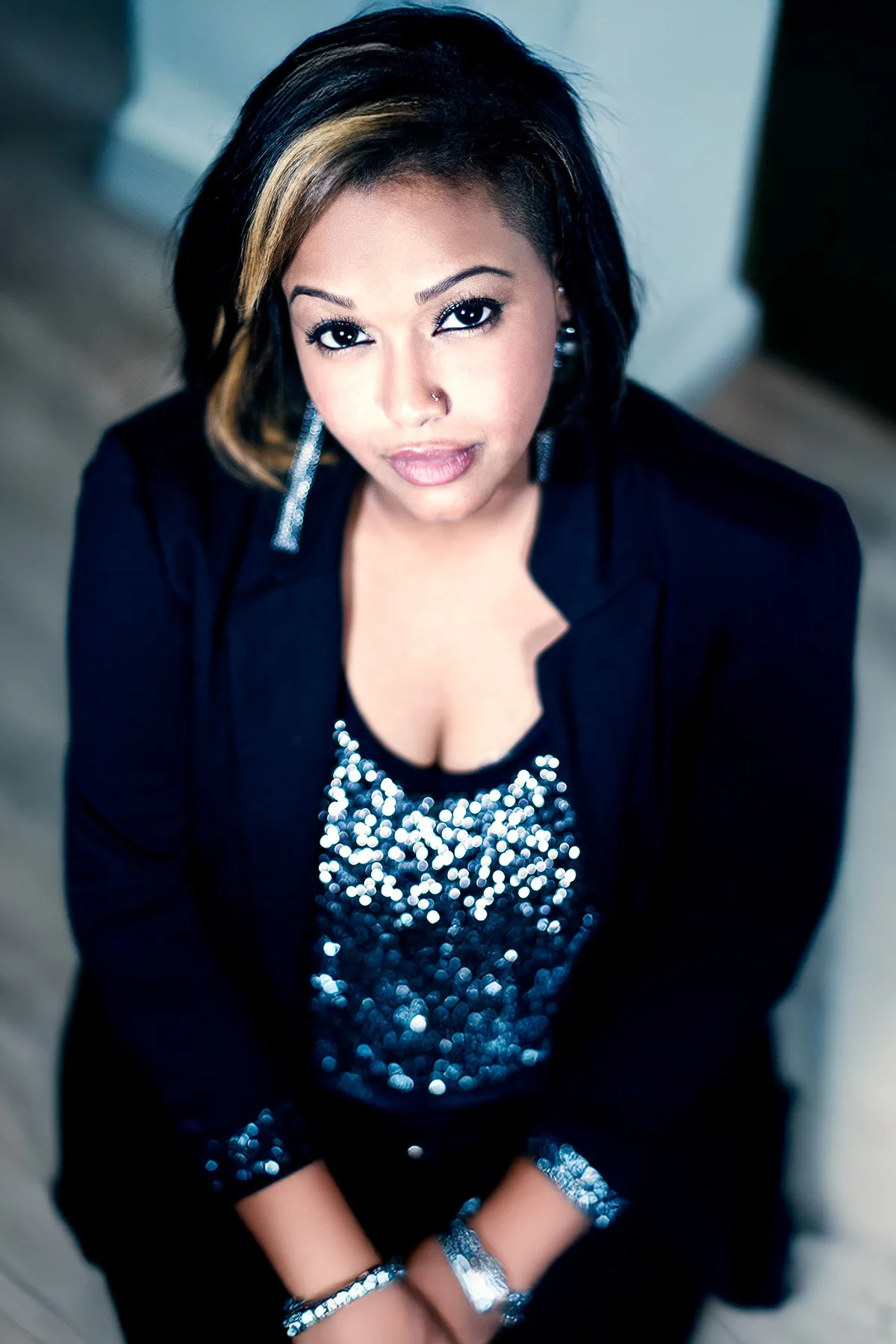 A woman with black and blonde hair, wearing a black blazer and a shiny sequin top, looking up at the camera.