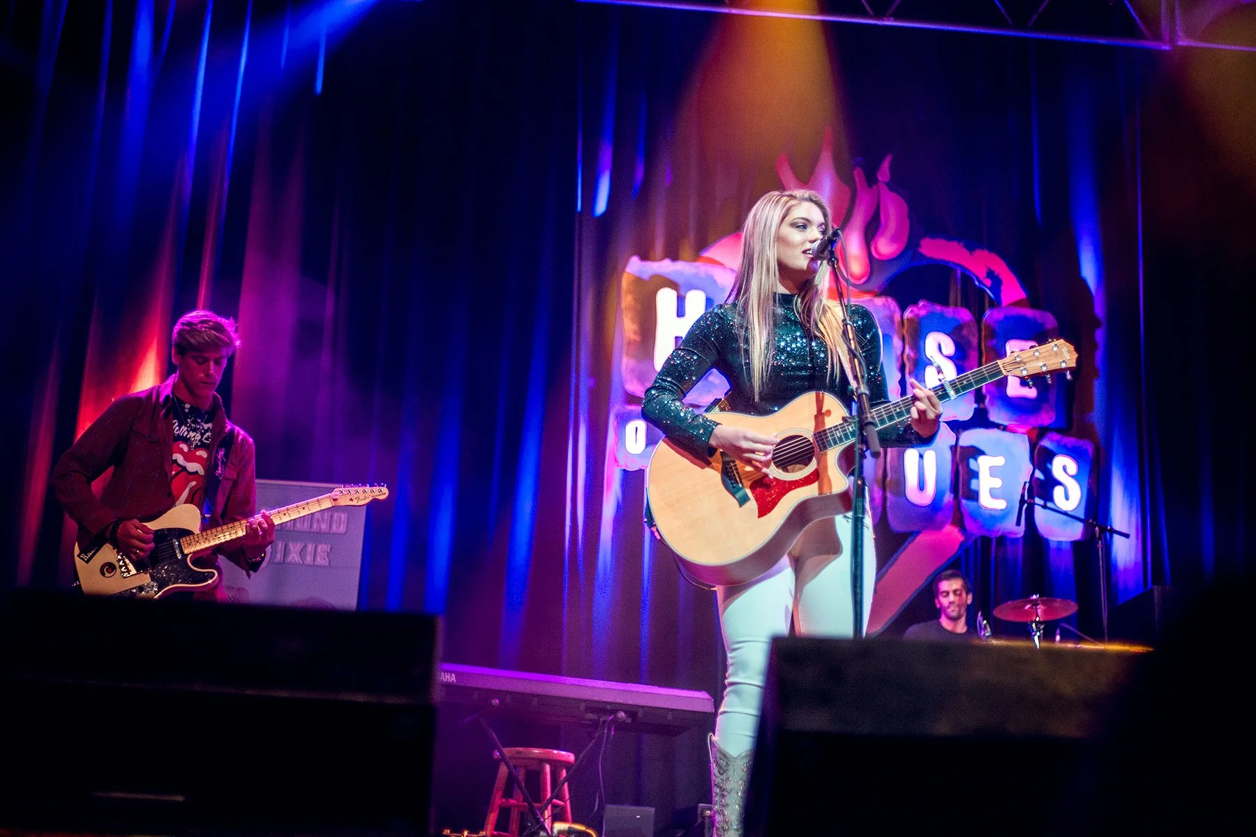 Female singer with long blonde hair performs on stage with acoustic guitar under colorful stage lighting, with male guitarist and drummer in the background at House of Blues.
