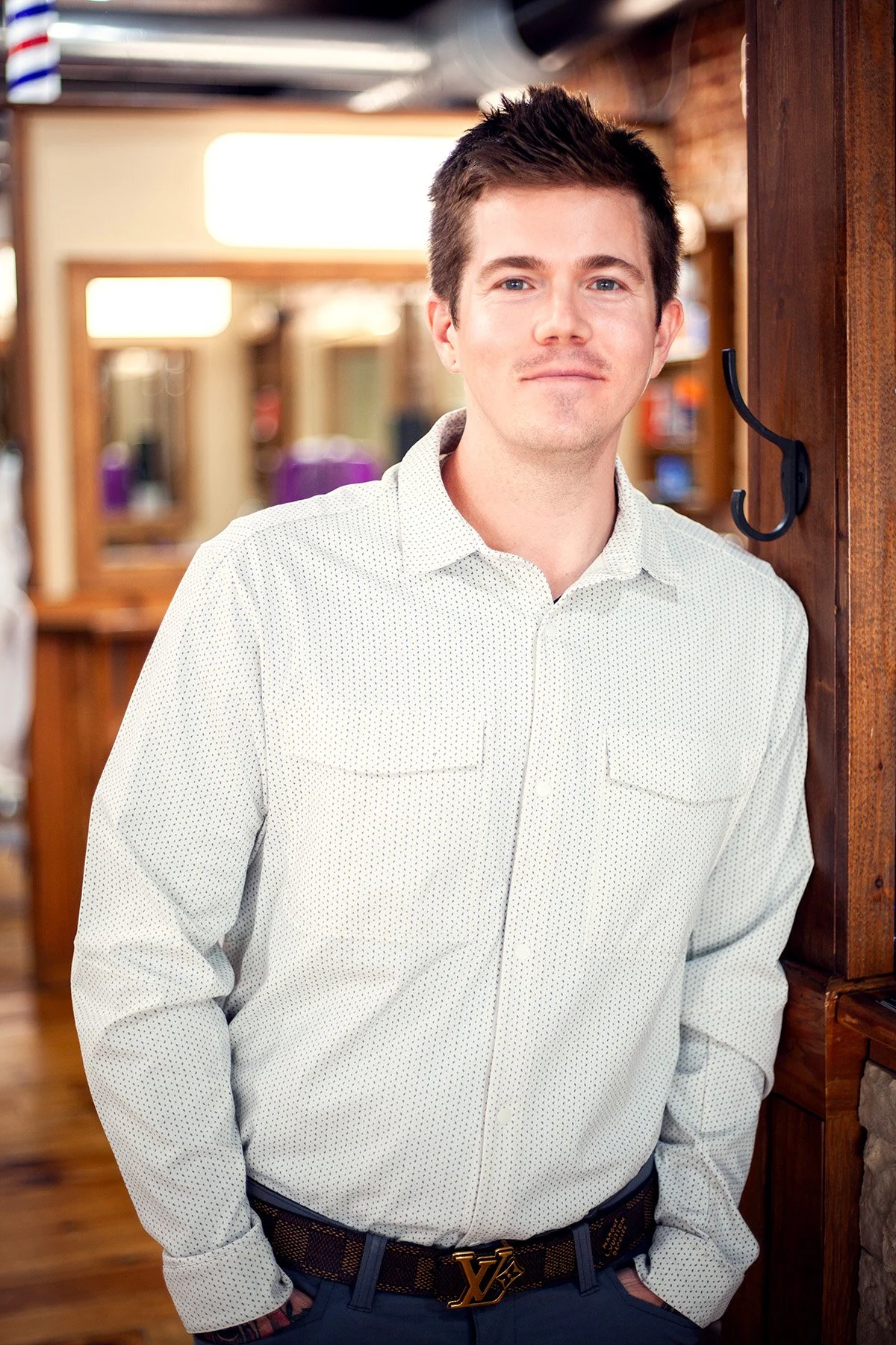A young man with short dark hair, blue eyes, and light skin, wearing a white button-up shirt with tiny black polka dots, standing indoors near a wooden wall hook with his hands in his pockets. The background is blurred with warm lighting and wooden d