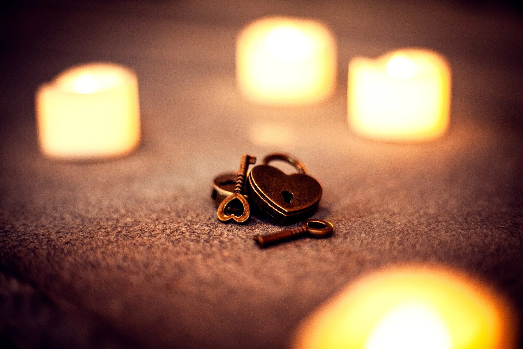 A small, dark heart-shaped lock with a tiny keyhole, surrounded by two miniature keys and a small heart-shaped key, all placed on a textured surface. The background features three softly blurred candle flames creating a warm glow.