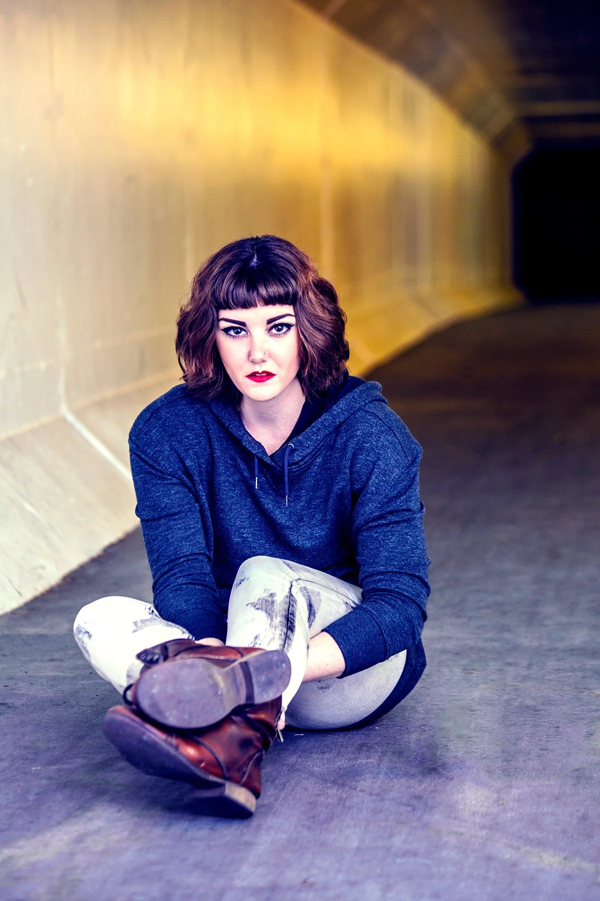 A young woman with dark, wavy hair and bangs, wearing a dark hoodie, white pants with black stripes, and brown boots, sitting on the ground in an underground tunnel or passageway with yellowish walls and a dark curved ceiling.