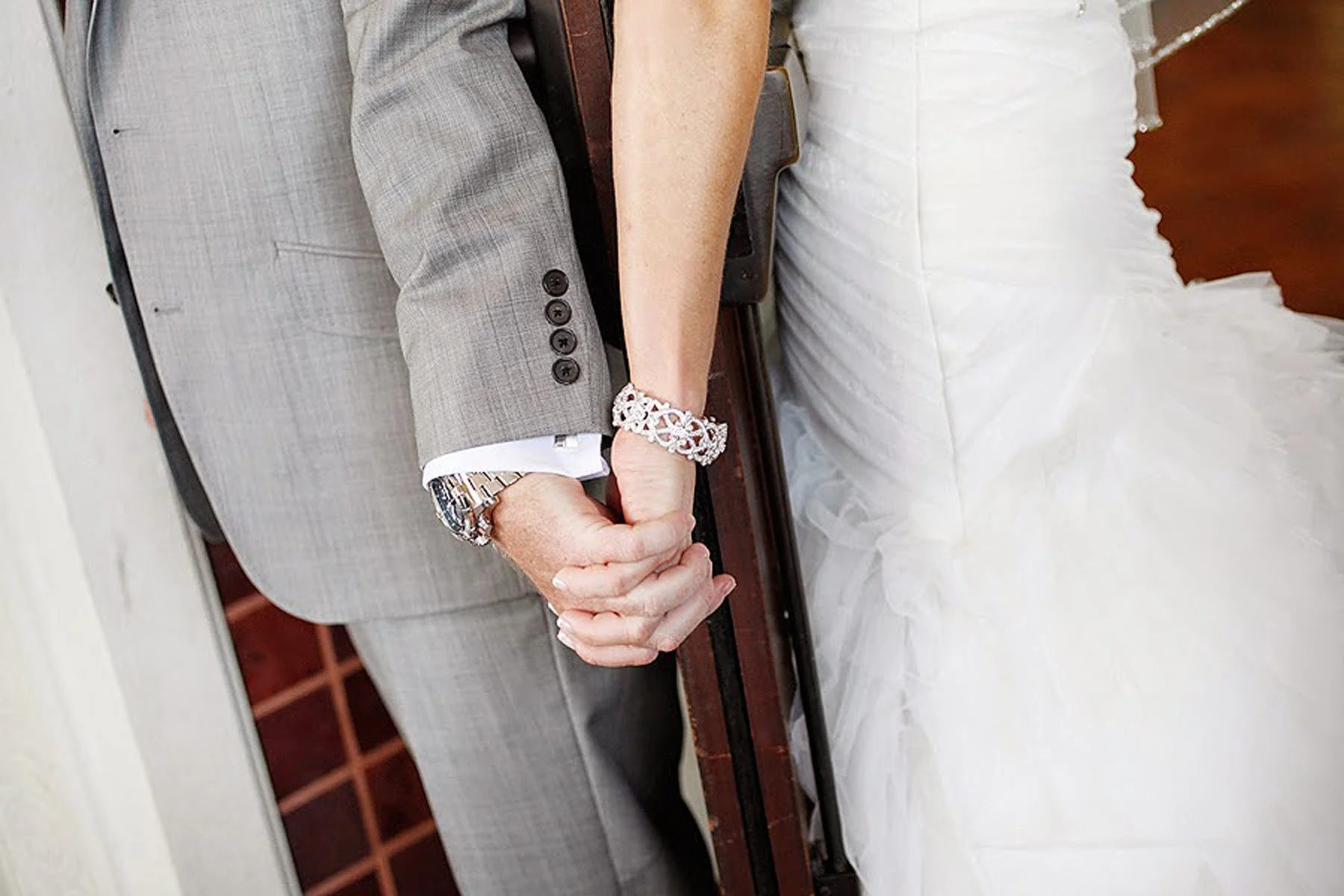 Close-up of a couple holding hands, wedding attire visible with the woman in a wedding dress and the man in a light gray suit with a watch and bracelet, against a background of a wedding setting.
