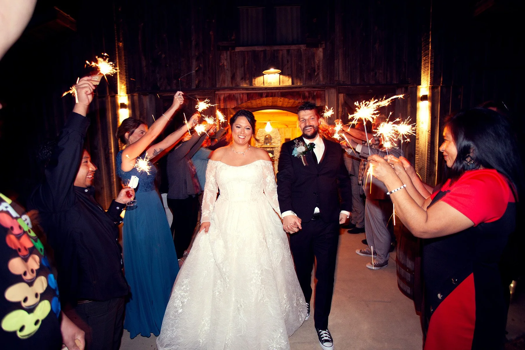 A bride and groom walking hand-in-hand through a corridor of guests holding sparklers, celebrating their wedding reception in a warmly lit, rustic venue.