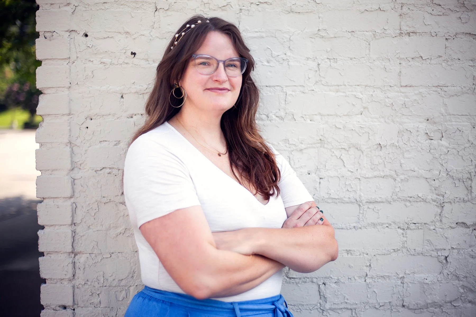 A woman with curly brown hair, wearing glasses, a white t-shirt, and blue pants, standing with arms crossed in front of a white brick wall.