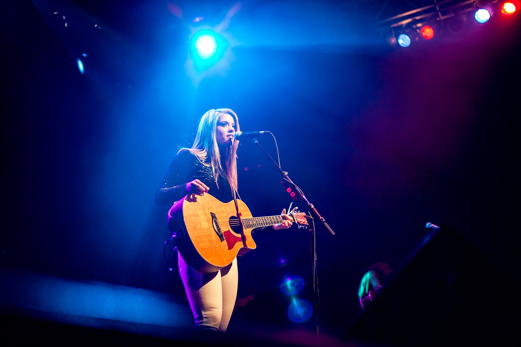 A young woman with long blonde hair playing an acoustic guitar and singing into a microphone on a stage illuminated with blue, purple, and red stage lights.