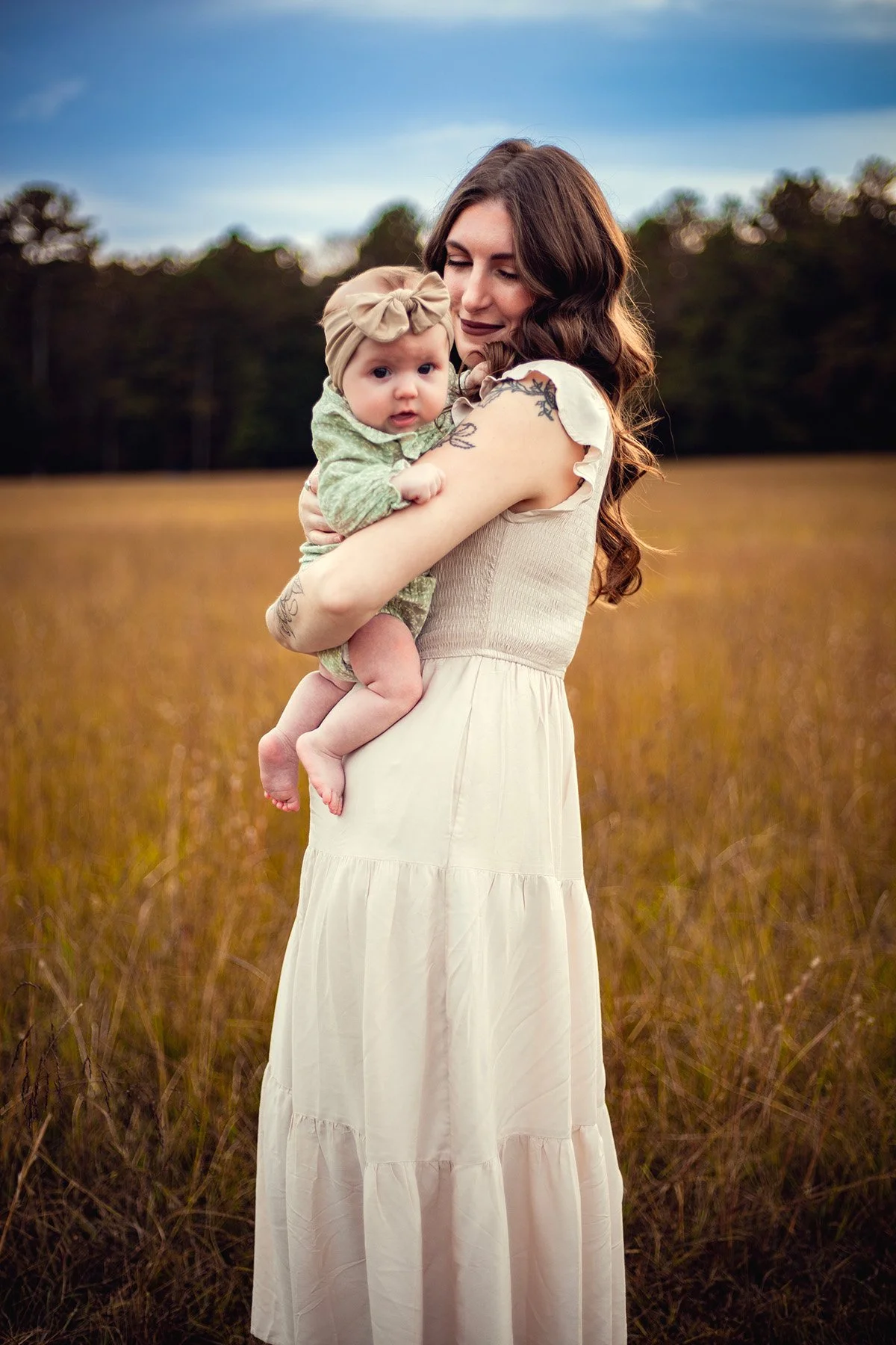 A woman with long brown hair, wearing a white dress, holding a baby in a field of tall grass with a background of trees and a partly cloudy sky.
