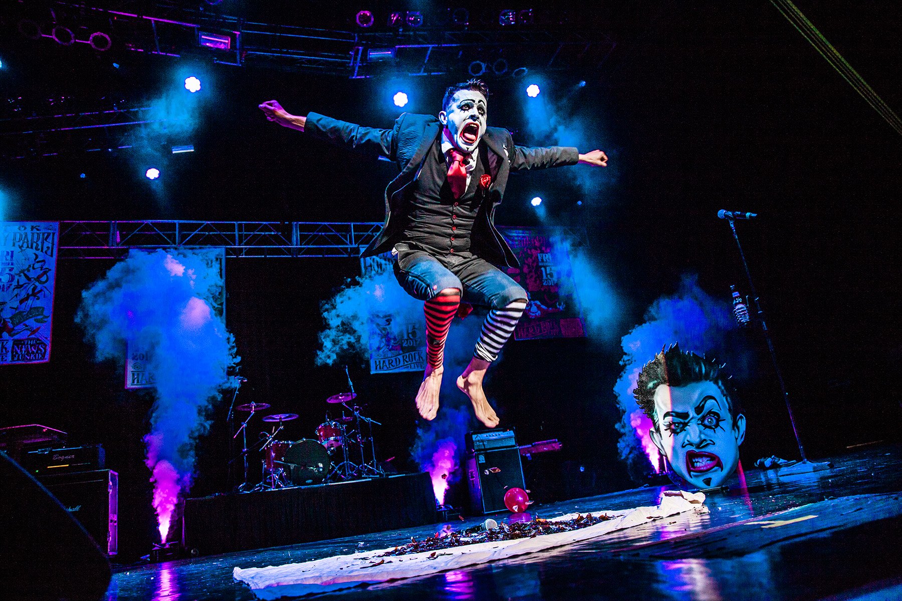 A performer mid-air on a stage, dressed as a clown with face paint, a black suit, striped red and black tights, and barefoot. The stage is lit with purple and blue lights, with smoke effects, drums, amplifiers, and oversized clown face decorations in