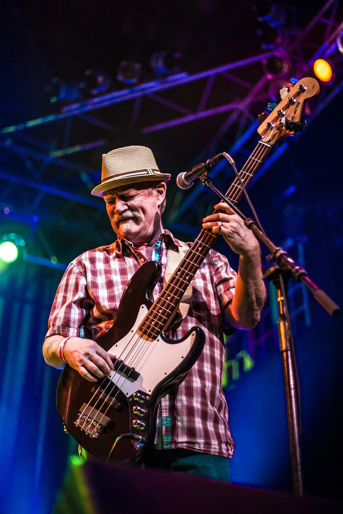 A man wearing a plaid shirt and a straw hat plays a bass guitar on stage with colorful lights and a metal framework above him.