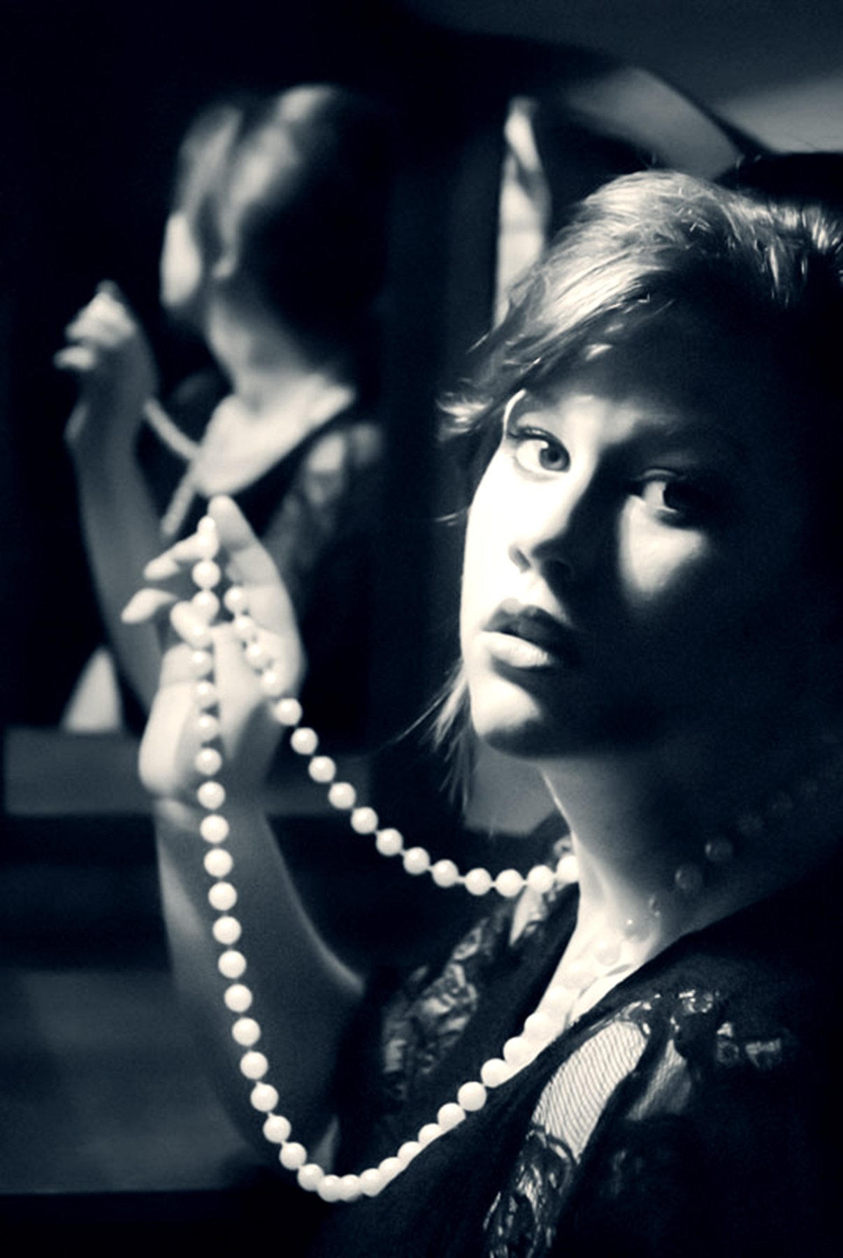 A black-and-white photo of a woman with wavy hair, holding a strand of pearl necklace, looking at the camera. Another woman in the background is slightly blurred, also holding a bead necklace.