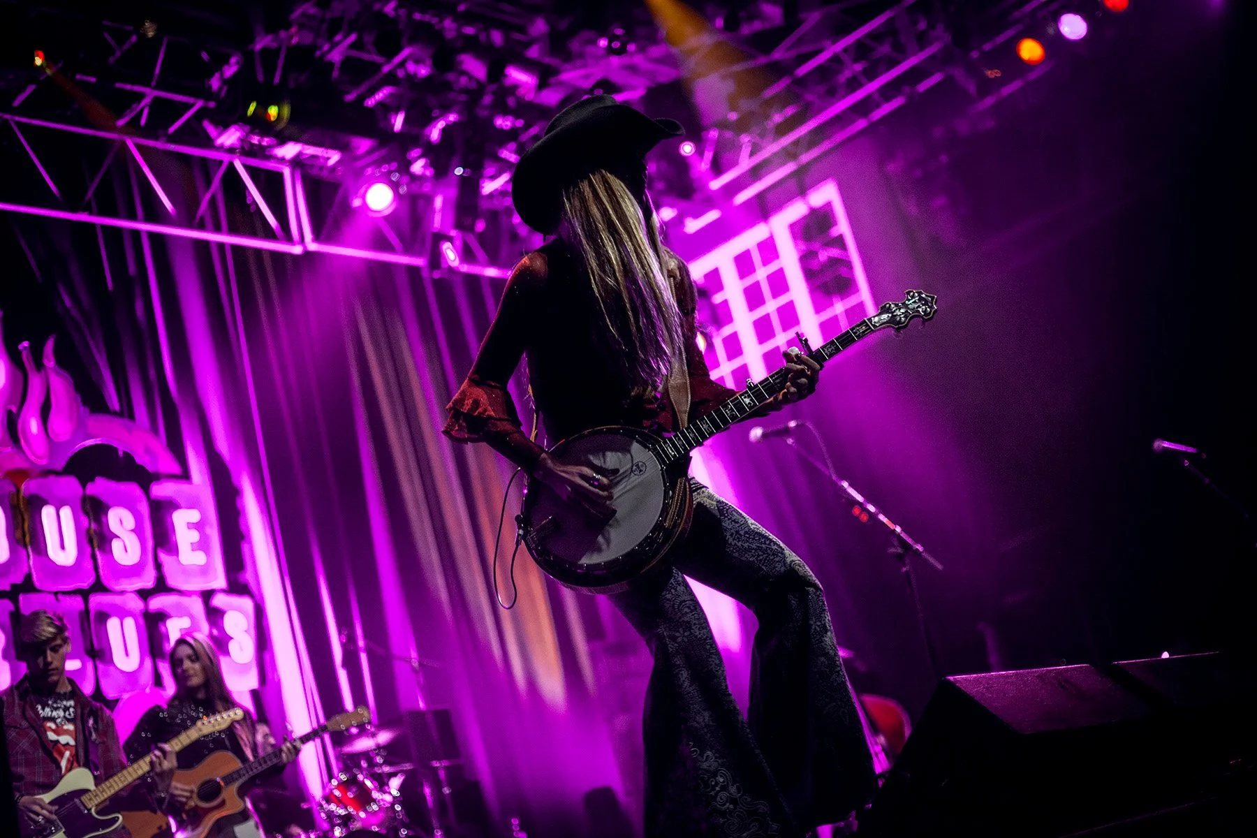 A woman playing an acoustic guitar on a stage illuminated with purple and pink lights, wearing a large black hat and patterned pants, with three other musicians in the background.