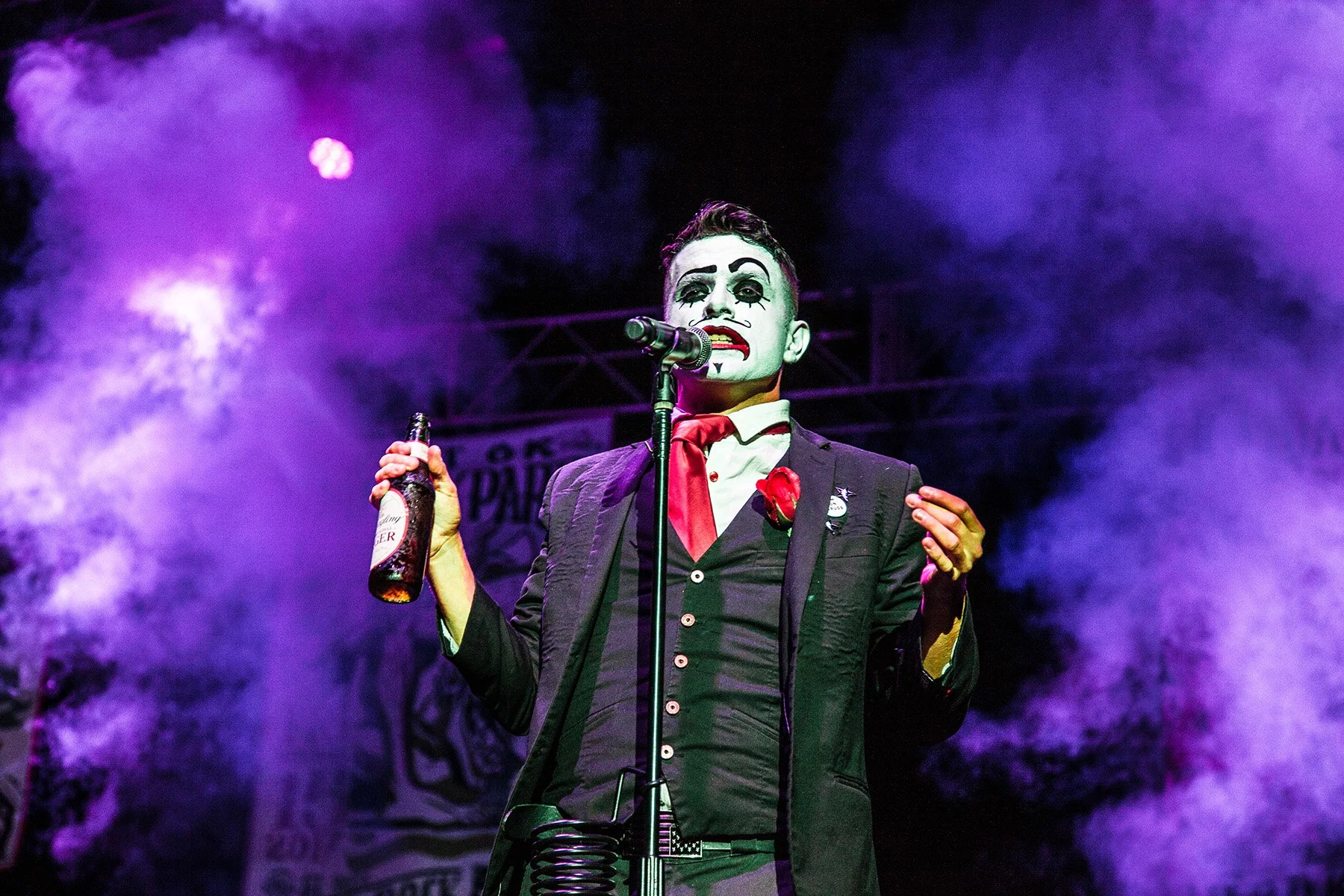 Performer dressed as a clown or mime with face makeup, holding a beer bottle, on stage with purple smoke and stage lighting in the background.