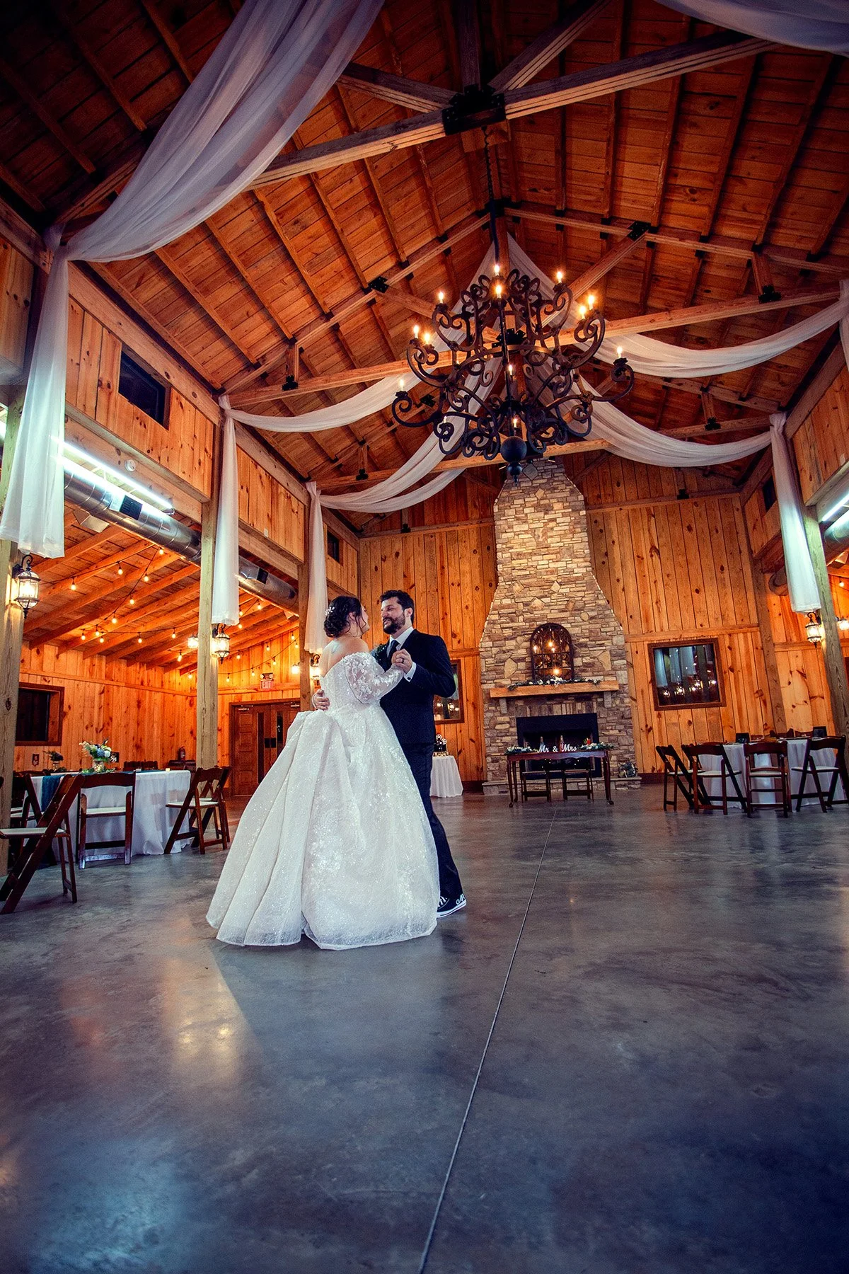 A bride and groom dancing in a rustic wooden wedding venue with a stone fireplace, chandelier, and draped white fabric on the ceiling.