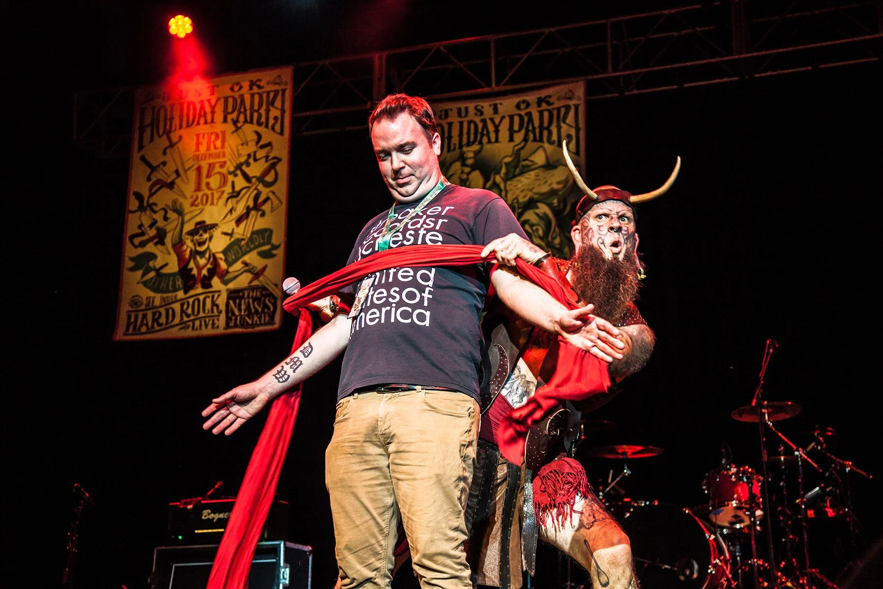 Two performers on stage at a concert, one dressed casually with a tattoo on his arm, and the other in a costume with a long beard, horned headpiece, and red attire. A backdrop displays the text "Holiday Party" with event details.
