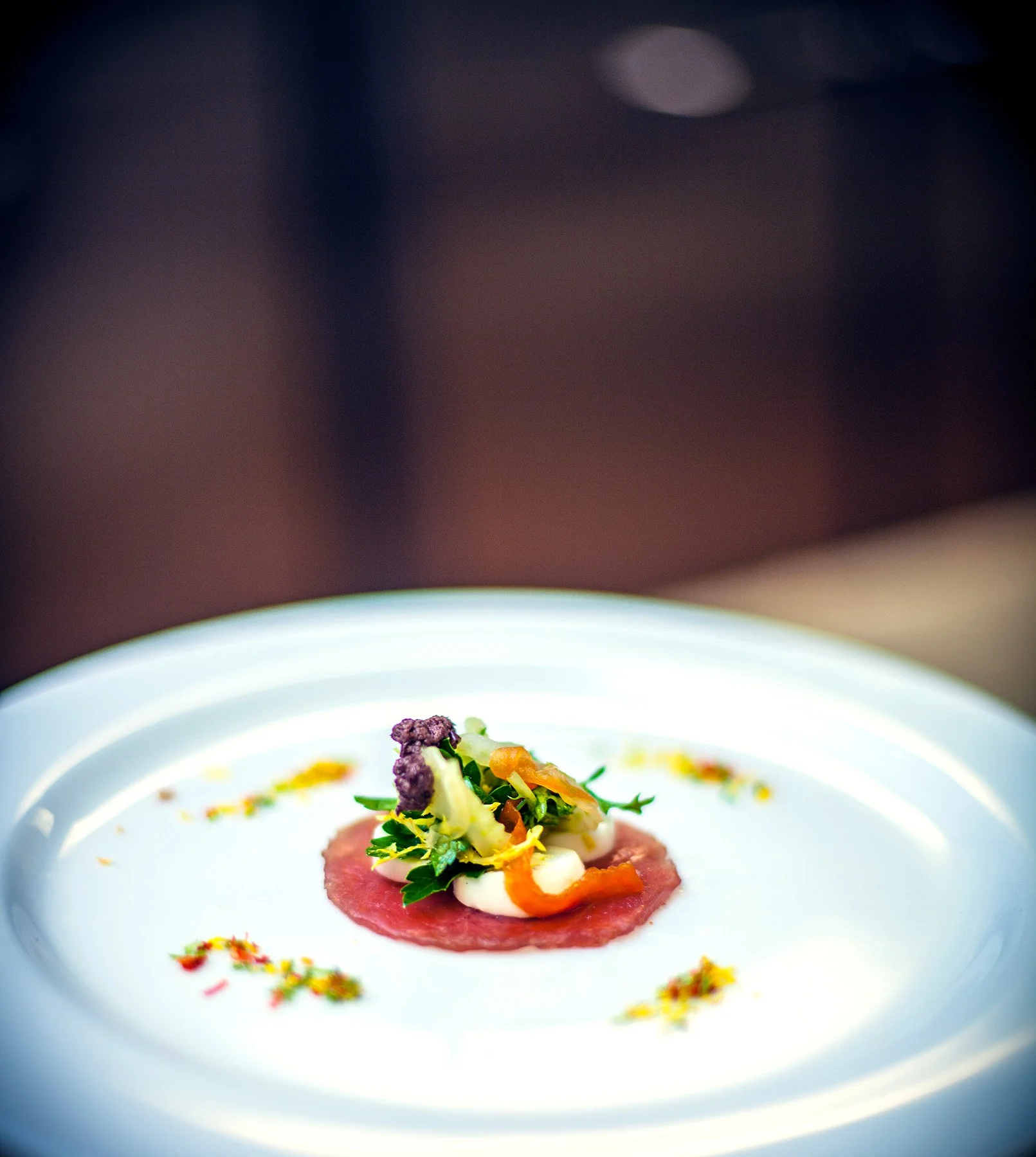 A plated gourmet dish featuring a thin slice of red meat topped with colorful vegetables and garnishes, arranged artistically on a white plate.