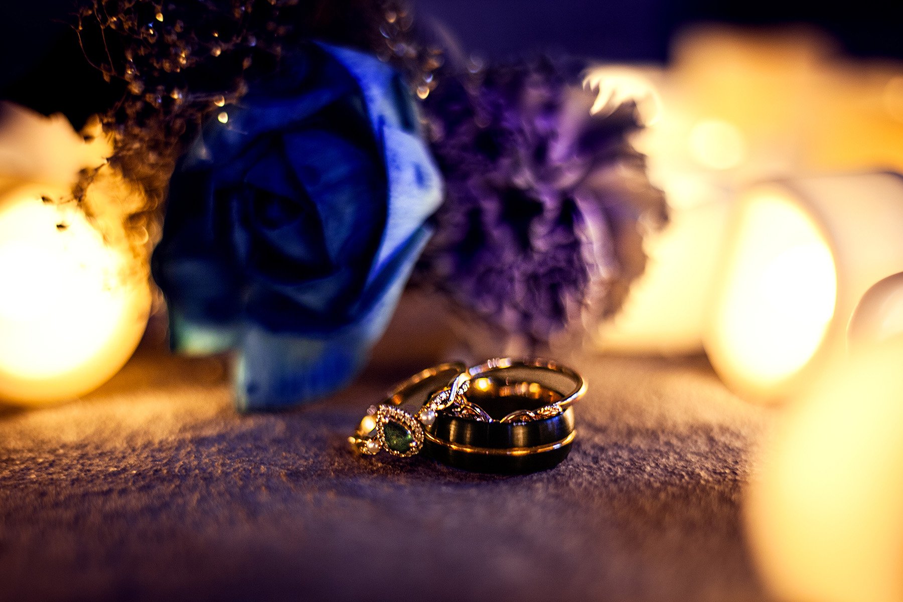 Close-up of wedding rings, including a gold engagement ring with a green gemstone and a black wedding band, placed on a surface with warm candlelight and blurred background of candles and a purple flower.