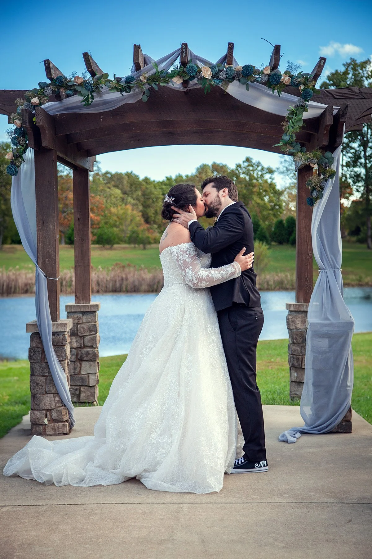 A newlywed couple sharing a kiss under an outdoor wedding arch decorated with flowers and white fabric, near a lake with trees and a blue sky in the background.