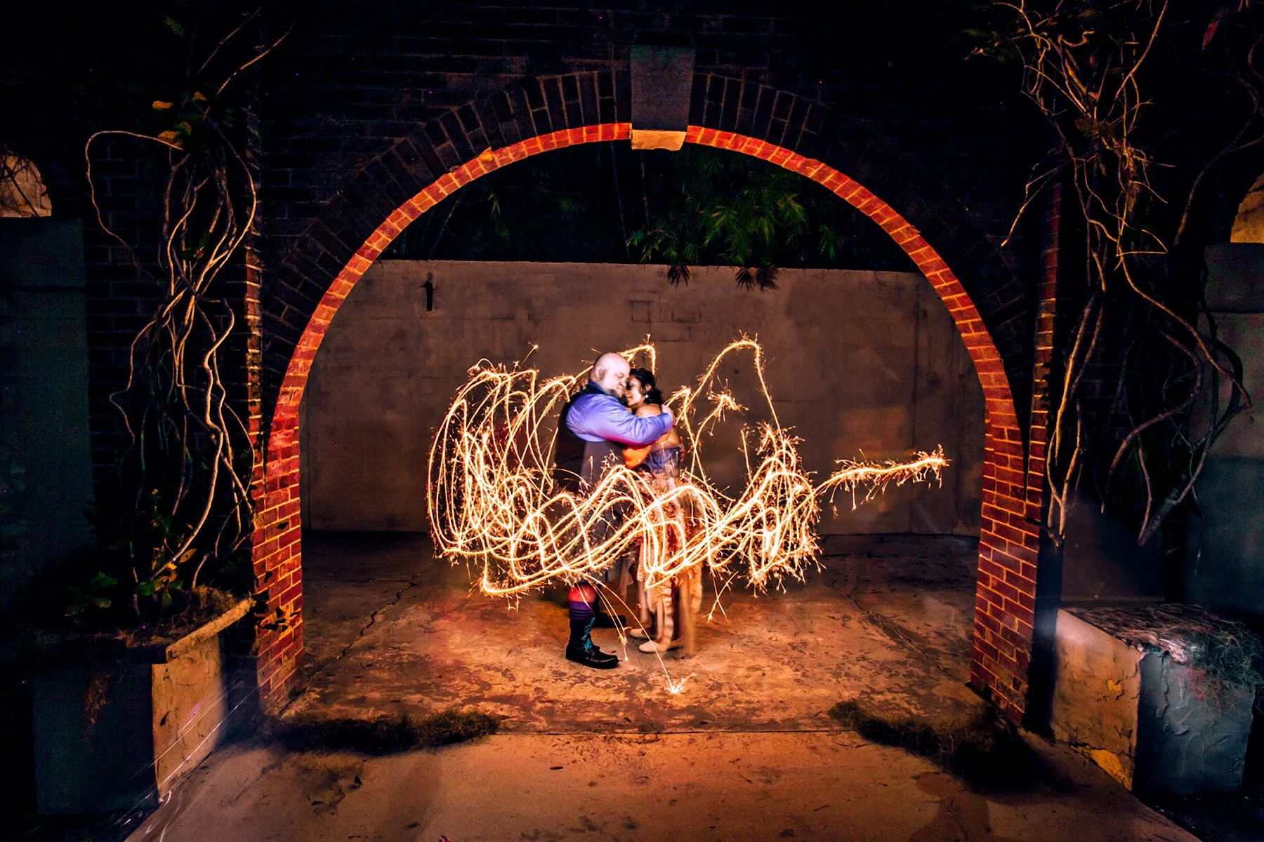 A couple dancing under an archway at night, surrounded by swirling sparklers creating a light trail effect.