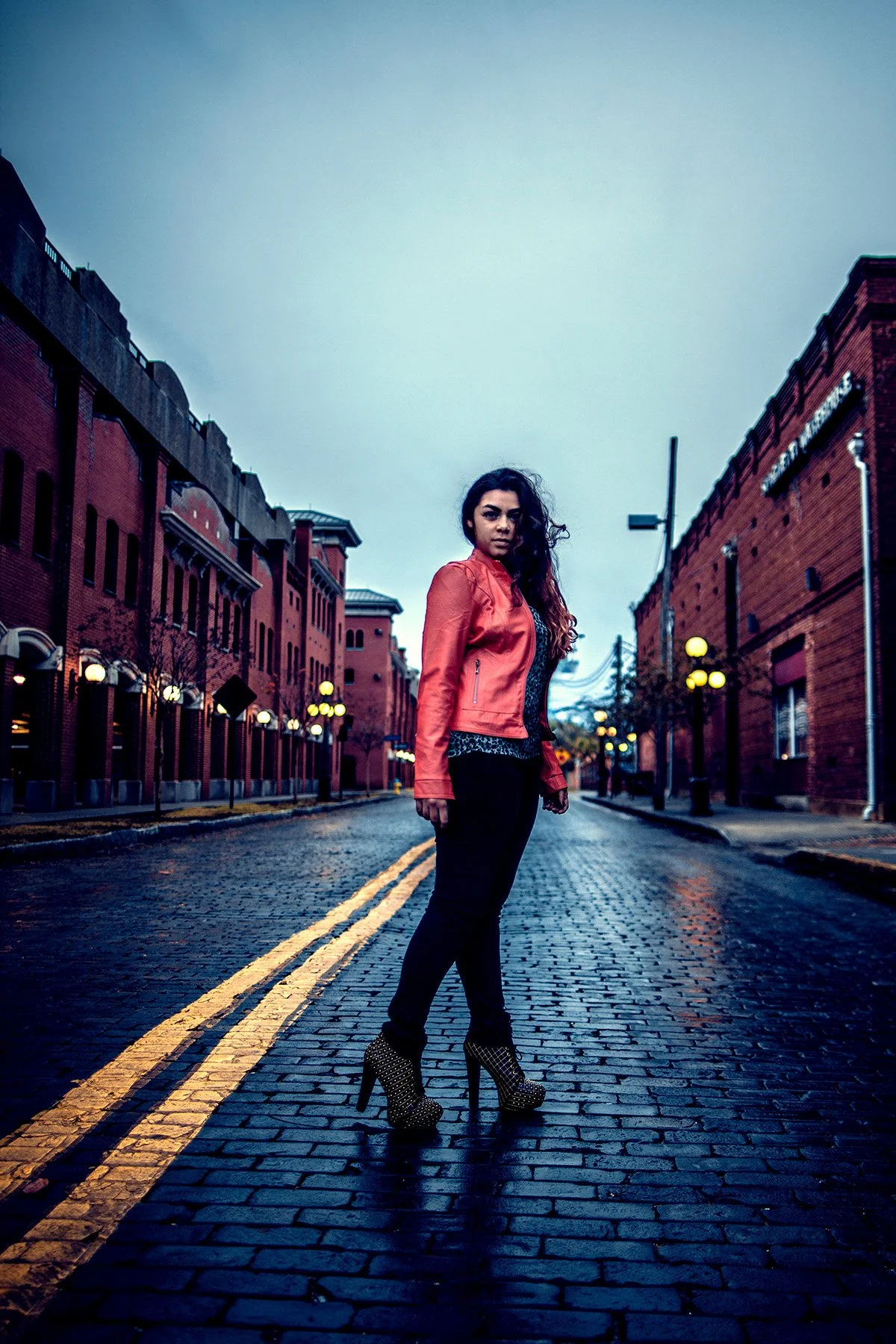 A woman with long curly hair stands on a wet cobblestone street in a city alleyway lined with brick buildings and street lamps, wearing a red leather jacket, black pants, and high-heeled shoes.