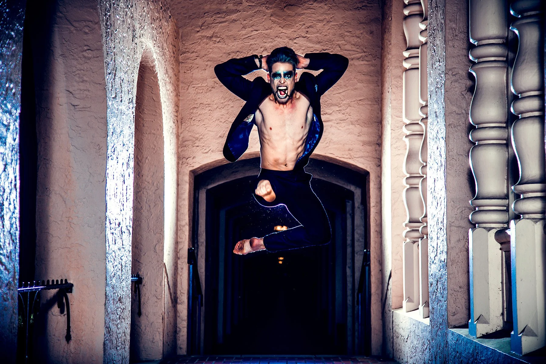 A man with dark hair, face makeup, and vampire fangs jumping mid-air in a narrow corridor with beige walls and decorative white columns. He is shirtless, wearing a dark jacket and black pants, with an intense expression.
