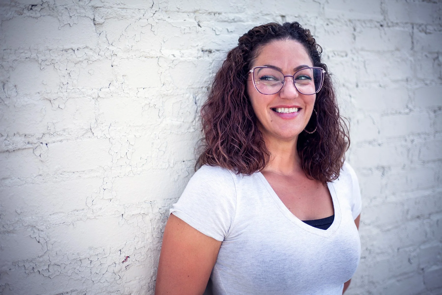 A smiling woman with curly brown hair and glasses leaning against a white, textured wall.