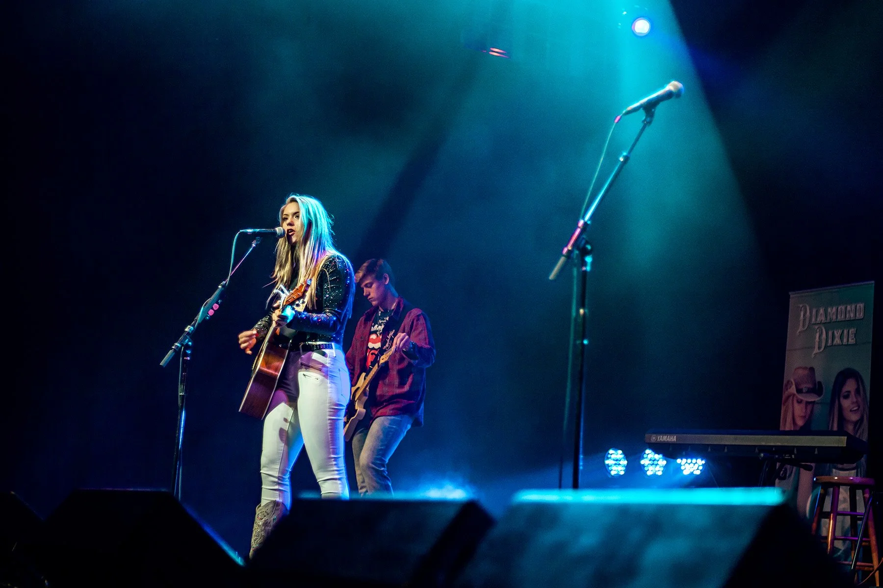 A female singer playing an acoustic guitar and singing on stage, with a male guitarist in the background, under blue stage lighting, in a concert setting.