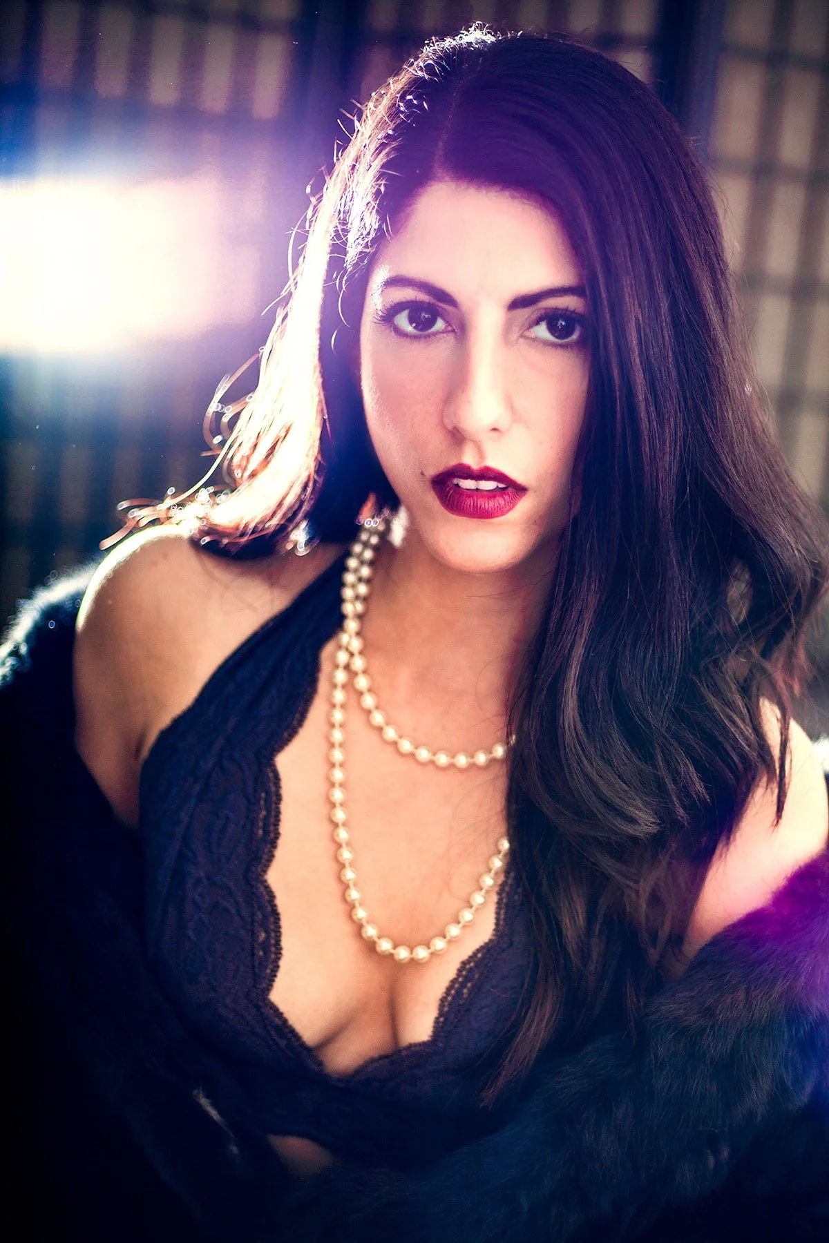 Portrait of a woman with long dark hair, wearing a blue lace dress, pearl necklace, and makeup with red lipstick, in a dimly lit setting with bright light behind her.