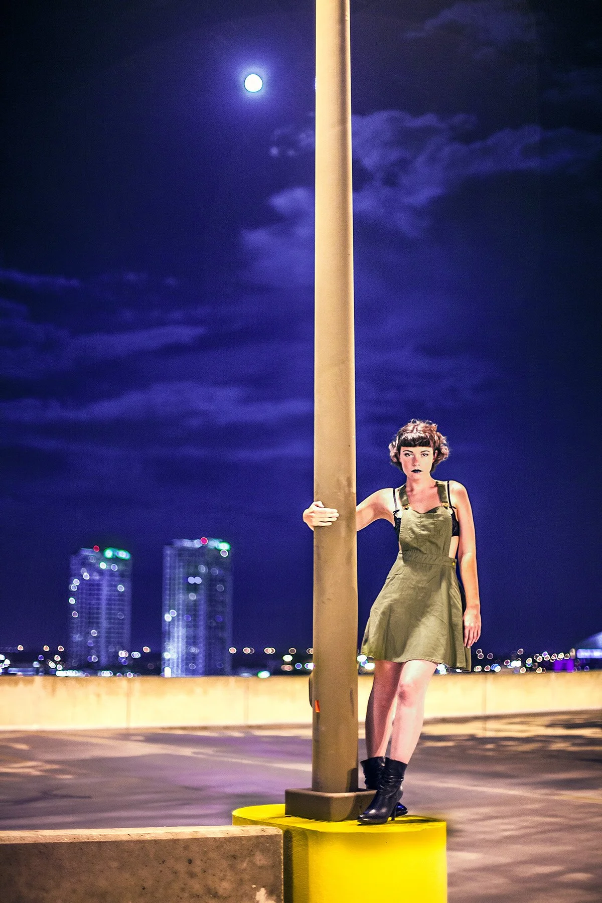 A woman with short dark hair, wearing a green dress and black boots, standing outdoors at night on a parking lot, holding onto a streetlight pole with a city skyline and moon in the background.