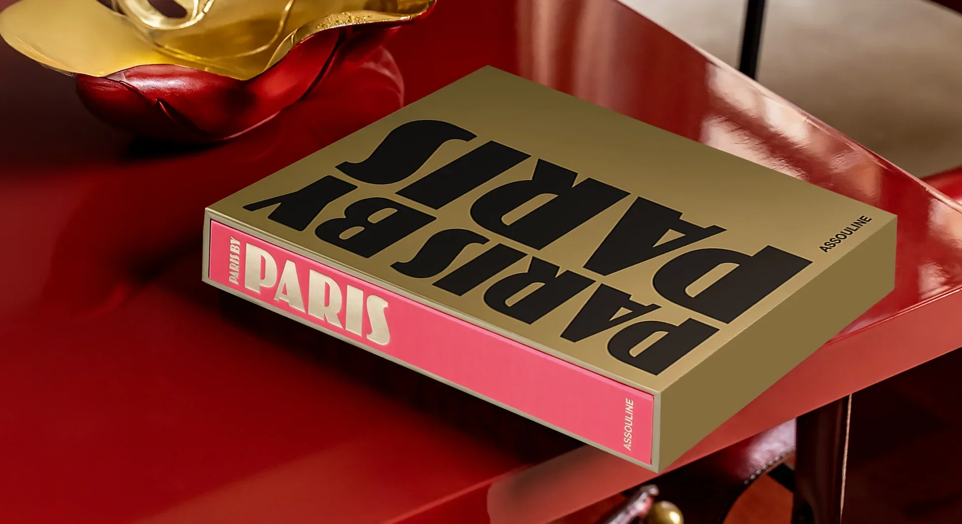 A book titled "Paris" by Assouline lies on a shiny red surface, with a partially visible decorative object above it. The book cover features large black text reading "PARIS" on a beige background with a pink spine.