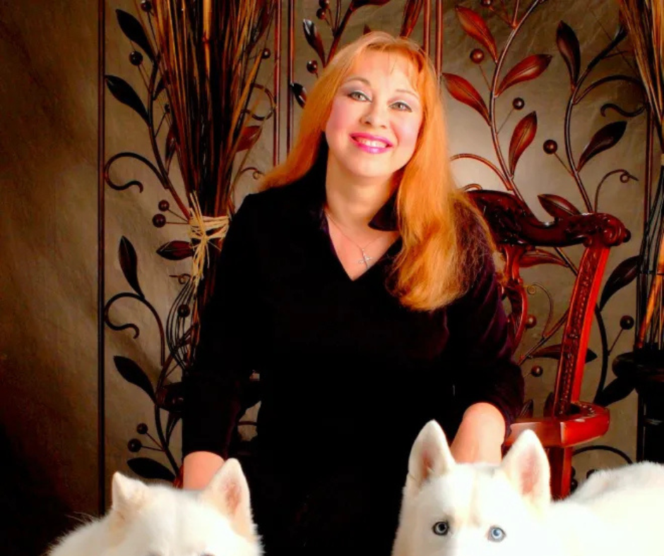 A woman with long, light orange hair sitting in front of decorative wooden panels with leaf and berry designs, smiling at the camera, with three white dogs near her, two of which have blue eyes.