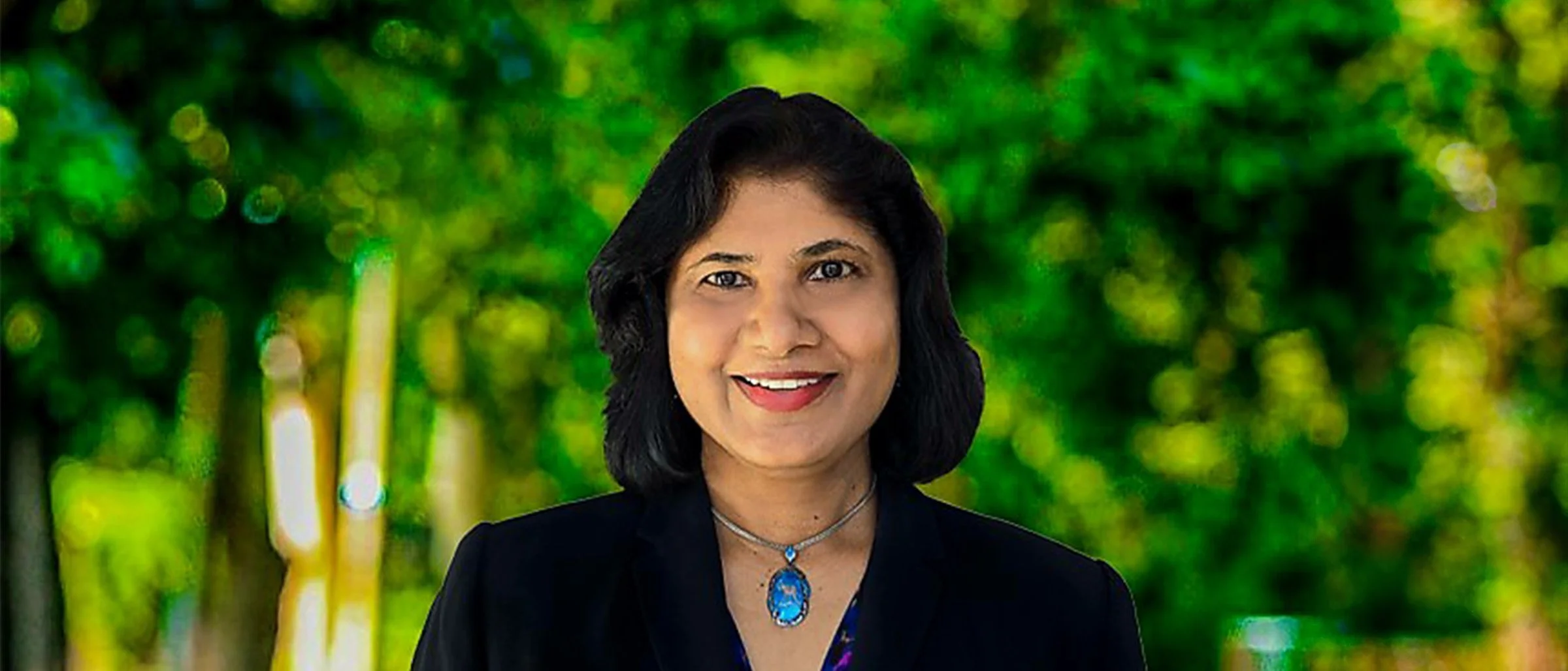 A smiling woman with shoulder-length black hair, wearing a black blazer and a blue gemstone necklace, standing outdoors with a blurred green foliage background.