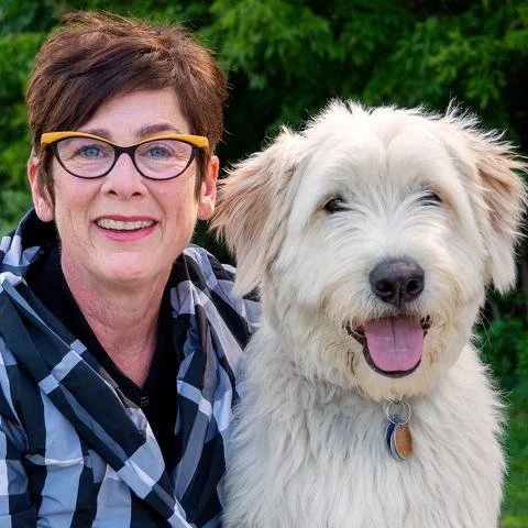 A woman with short brown hair, glasses, and a plaid shirt standing next to a white, fluffy dog with its tongue out, outdoors with green foliage in the background.
