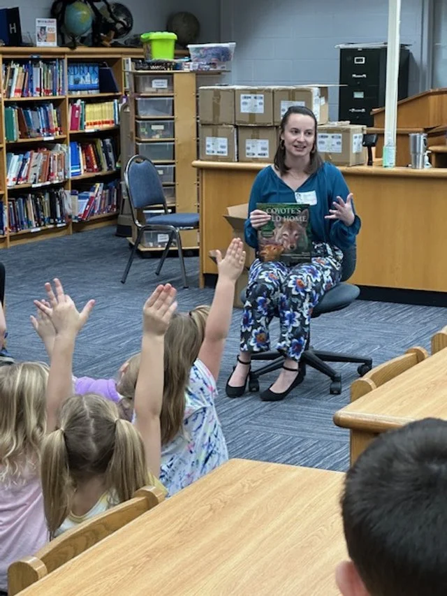 A woman reading to a group of young children in a library or classroom setting, with children raising their hands.