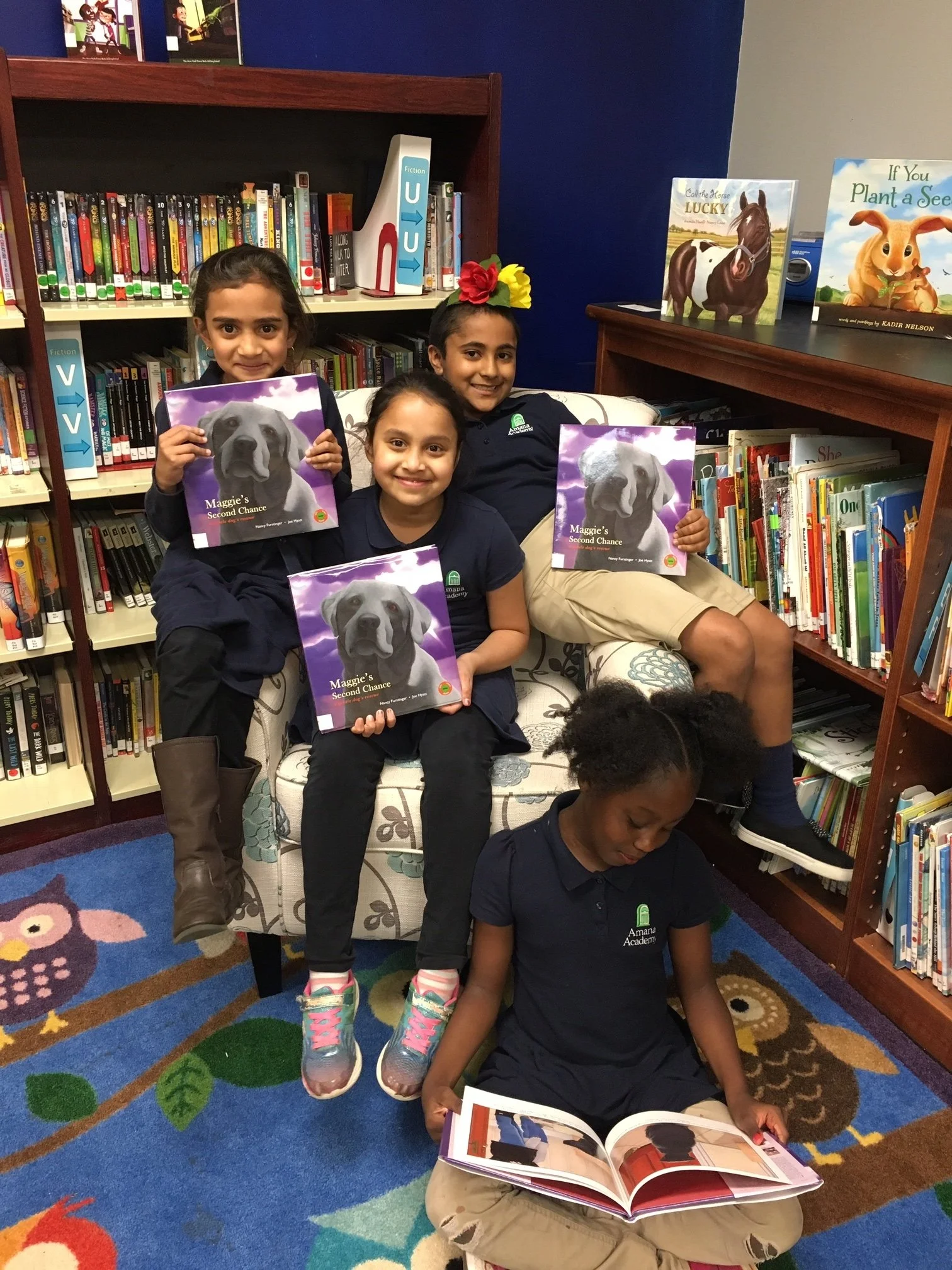 Four girls in school uniforms in a library, three sitting on a couch holding books titled 'Maggie's Second Chance' with a Labrador retriever on the cover, one girl sitting on the floor reading a book, all smiling and surrounded by bookshelves with ch