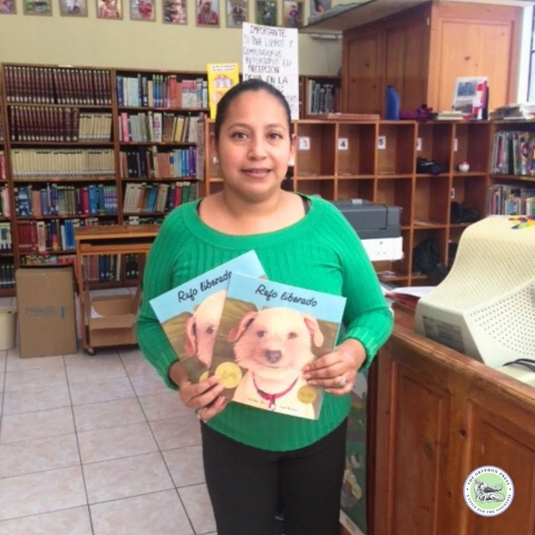 A woman standing in a library holding two copies of a book titled "Rufo liberado" featuring a puppy on the cover. She is wearing a green sweater and has tied hair back. Bookshelves and cubbyholes are visible in the background.