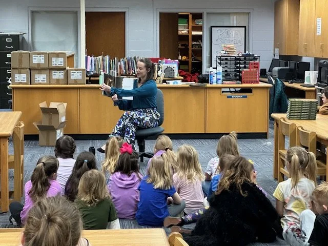A woman reading a book to a group of young children seated on the floor in a library classroom.